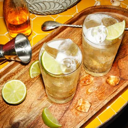Gin Buck in two highball glasses on a wooden tray with ice, lime wedges and a metal jigger