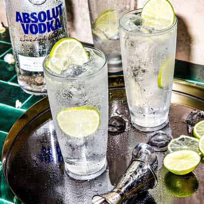 Hard Seltzer Drink in two highball glasses with ice and lime slices beside an Absolut Vodka bottle on tiles