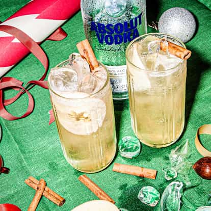 Very festive table with two Holiday Mule cocktails and a bottle of Absolut Vodka, stand beside Christmas paper, streamers, cinnamon sticks and apple slices.