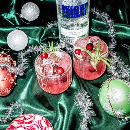 Very Christmassy table with two Jingle Buck cocktails on a green velvet table cloth stand beside a bottle of Absolut Vodka and decorative Christmas balls and tinsel.
