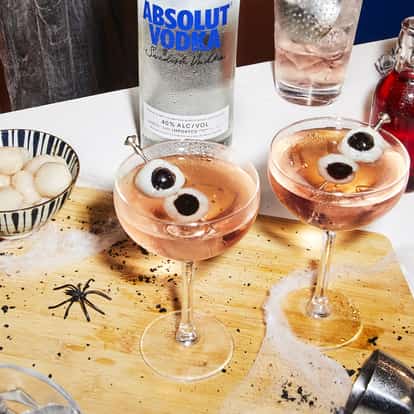 Lychee Eyeball Martini in coupe glasses with skewered lychees on a wooden board with an Absolut bottle behind