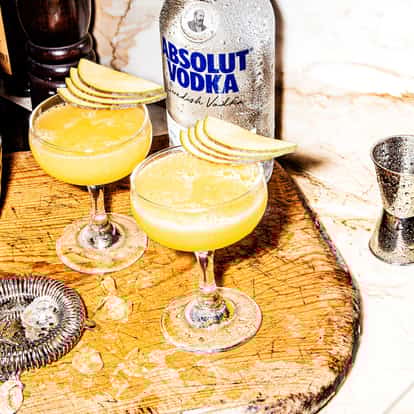 Maartens Mango Martini in two coupe glasses garnished with apple slices and an Absolut Vodka bottle behind