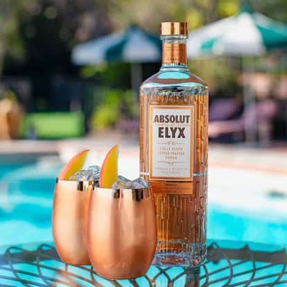 Elyx ManGo two metal tumblers with ice and mango wedges beside a vodka bottle on a glass table by a pool