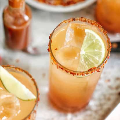 Michelada in two tall glasses rimmed with chili salt and garnished with lime wedges and ice