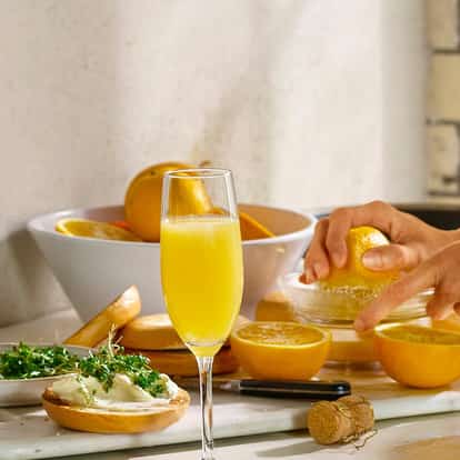 Mimosa cocktail next to a bottle of champagne surrounded by fresh oranges.