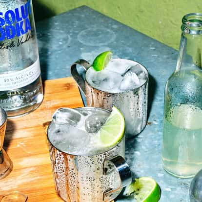 Two Moscow Mule cocktails in two metal Mule Mugs next to a bottle of Absolut Vodka.