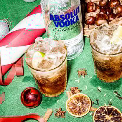 Two Mulled Mule cocktails on a Christmas-themed table with Christmas wrap and a bottle of Absolut Vodka laying nearby.