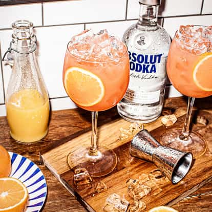 Orang-a-tang two wine glasses filled with crushed ice and orange wheel garnishes, Absolut Vodka bottle on a wooden board