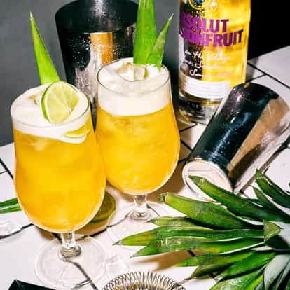 Passion For Days in two stemmed glasses with lime wheels and pineapple leaves, Absolut bottle on tiled table