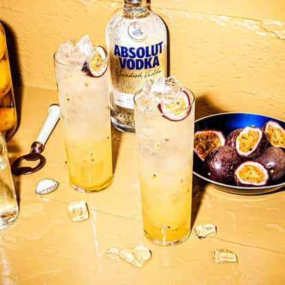 Passion Fruit Collins in two Collins glasses with ice and halved passion fruit, Absolut vodka bottle on table