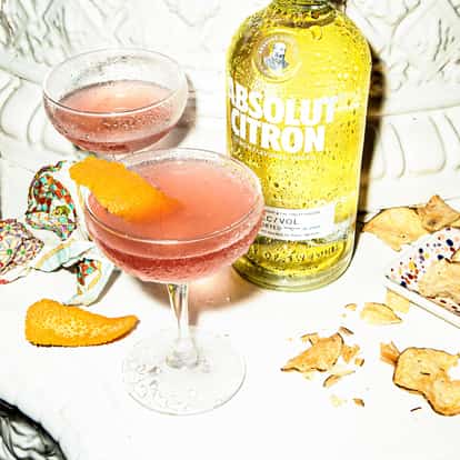 Pomegranate Martini in a coupe glass with an orange twist and an Absolut Citron bottle on a white surface
