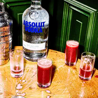 Absolut vodka bottle beside five red-purple cocktails in highball glasses on a wooden table.