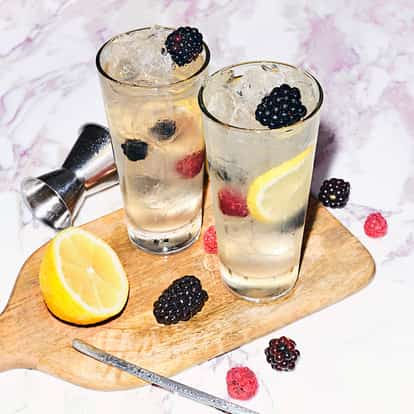 Ragnar 2.0 in two highball glasses with crushed ice a lemon slice and blackberries and raspberries on a wooden board
