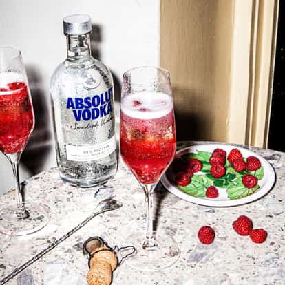 Raspberry Bellini served in two champagne flutes with raspberries and an Absolut Vodka bottle on a marble table