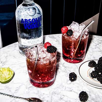 Two raspberry cocktails in rocks glasses with crushed ice and blackberries, a bottle of Absolut vodka on marble table