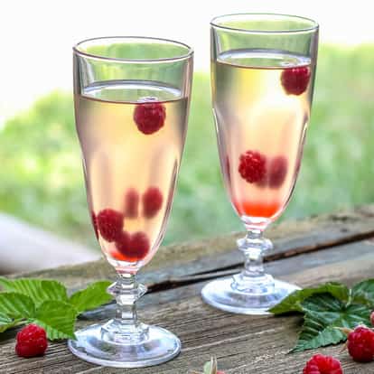 Raspberry Cocktail in two tall fluted glasses with whole raspberries and raspberry leaves on a wooden table