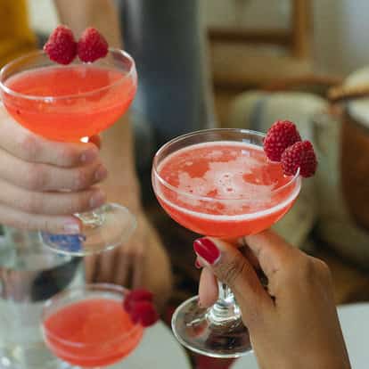 Raspberry Martini served in coupe glasses garnished with raspberries held by two hands