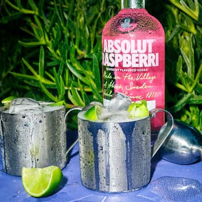 Two Raspberry Mule cocktails in stainless steal Mule Mugs next to a bottle of Absolut Raspberri set against a background of green leaves.