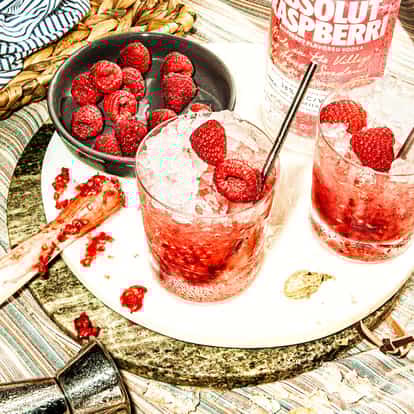 Two delicious-looking raspiroska cocktails next to a bottle of Absolut Raspberri and fresh raspberries.