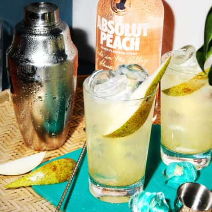 Sapodill Fizz served in two highball glasses with pear slices and ice, Absolut Peach bottle visible