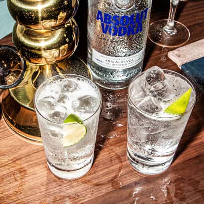 Skinny Bitch in two highball glasses with ice and lime wedges and an Absolut vodka bottle on a wooden table