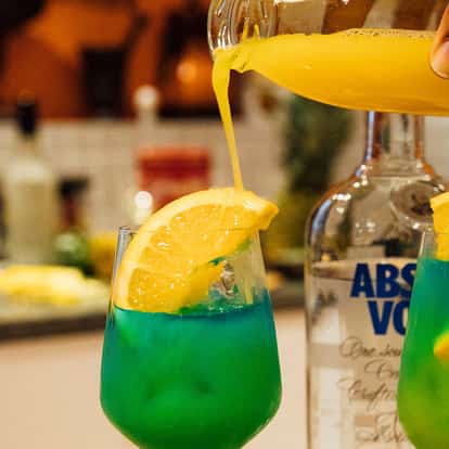 Skylab in two stemless glasses with lemon wedges and a hand pouring orange juice, Absolut bottle behind