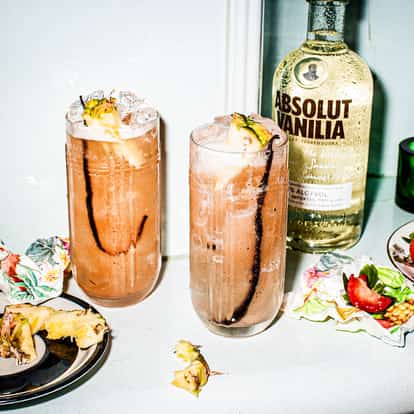Two long glaases of strawberry night cocktail on a table with Absolut Vanilia bottle and plates with sliced pine apple and strawberries.