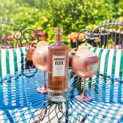 The Elyx Spritz with Absolut Elyx bottle between two stemmed glasses of crushed ice and lemon on a patio table