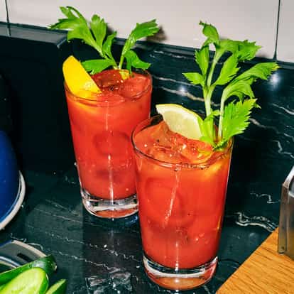 Virgin Mary in two highball glasses with celery stalks and lemon wedges, ice and pepper on countertop