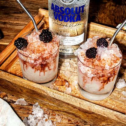 Vodka Bramble in two rocks glasses with crushed ice and blackberries and Absolut Vodka bottle on a wooden tray