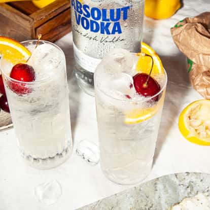 Vodka Collins in two highball glasses with orange wedge and cherry and an Absolut Vodka bottle in the background