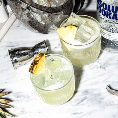 Vodka Fizz in two rocks glasses with pineapple wedges and an Absolut bottle on marble background