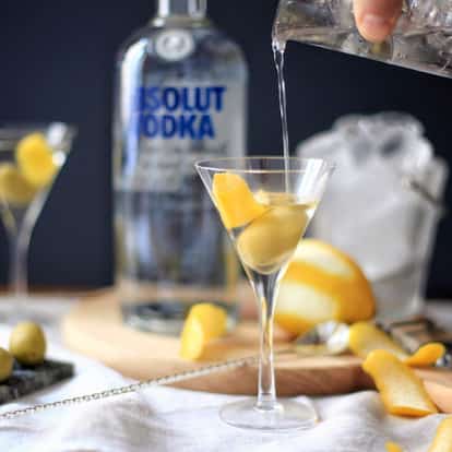 Vodkatini in a martini glass with olives and a lemon twist and an Absolut vodka bottle in the background