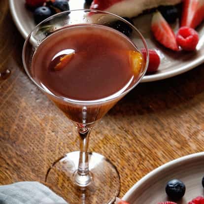 Volga Boatman in a stemmed cocktail glass with an orange peel, on a wooden table with plates of berries.