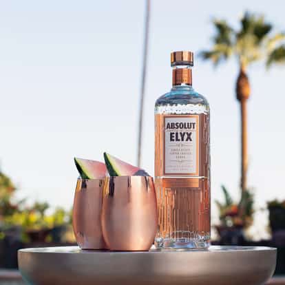 Absolut Elyx vodka bottle next to two copper mugs with watermelon wedges on a low round tray.