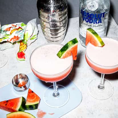 Two Watermelon Martini cocktails beside a bottle of Absolut Vodka on a glass table.