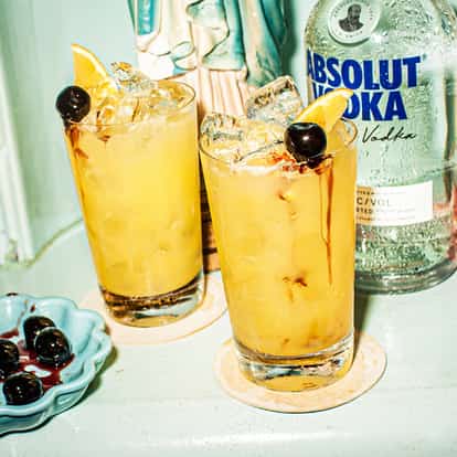 Yellowhammer Cocktail in two highball glasses with ice and lemon wedge and cherry garnish, Absolut Vodka bottle at right