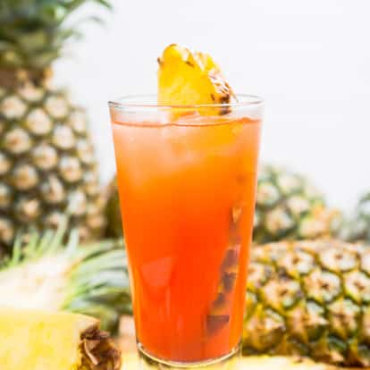 Zombie in a highball glass with pineapple wedge garnish on a wooden board with pineapple halves in background