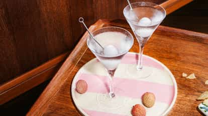 Two chilled lychee martinis in martini glasses with skewered lychees, served on a striped tray with fresh lychees.
