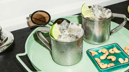 Cold metal mugs filled with ice and lime wedges, served on a green tray with a side of cashew nuts and sunglasses in the background.