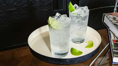 Dos vasos con un cóctel claro y burbujeante con hielo y gajos de lima, servidos en una bandeja redonda blanca con más trozos de lima al lado. El entorno es moderno, con libros y un fondo oscuro.