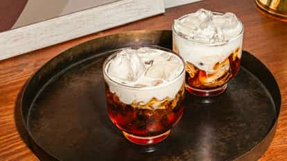 Dos vasos bajos con un cóctel en capas de licor oscuro, hielo y una cubierta blanca y cremosa, servidos en una bandeja oscura sobre una superficie de madera. La bebida se ve fría y de textura rica.