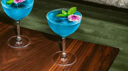Lotus Flower Martini cocktail served in a coupe glass with a bright blue color, garnished with a pink flower and fresh mint, on a wooden table.