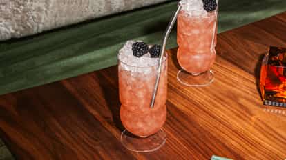 Cóctel A Veranda Moment en vaso alto con hielo picado, moras y popote metálico, de pie sobre una mesa de madera.