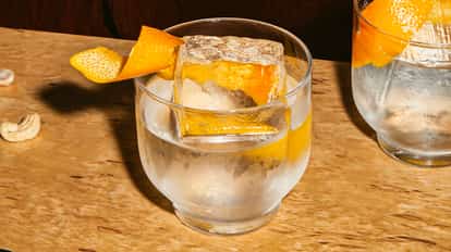 Chilled Absolut Gangster cocktail in a round glass with a large clear ice cube and orange peel garnish on a wooden table, with cashews scattered nearby.