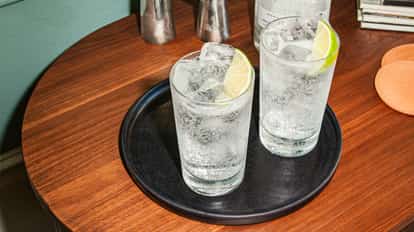 Two vodka soda cocktails with ice cubes and lime wedges served in highball glasses on a black tray on a wooden table.