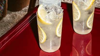 Zwei hohe Gläser Vodka-Limonade mit Eiswürfeln und Zitronenscheiben auf glänzend rotem Tisch, erfrischender Zitrus-Cocktail.