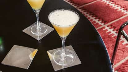 Absolutely Bananas cocktails served in chilled martini glasses with a light, frothy top, set on a dark table with clear square coasters.