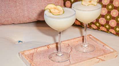 Two Apple Strudel Martini cocktails in coupe glasses with dried apple slices on a pink marble tray, set on a soft sofa with patterned pillows in the background.