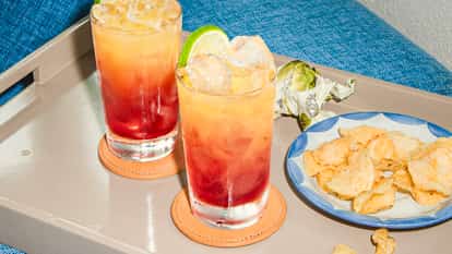 Two Bay of Passion cocktails with red-to-orange layers and lime wedges on a tray beside chips, set against a blue sofa.